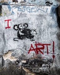 A piece of street art on a textured wall featuring a stencil of a monkey holding a paint roller and a rat. The words 'I ART!' are painted in bold red letters beside the figures.