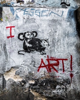 A piece of street art on a textured wall featuring a stencil of a monkey holding a paint roller and a rat. The words 'I ART!' are painted in bold red letters beside the figures.