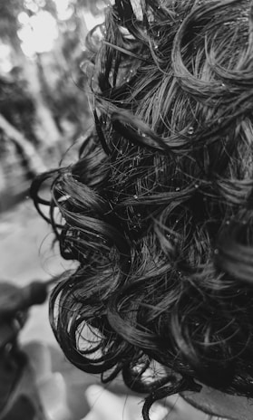 Close-up view of dark, curly hair with visible texture and moisture. The curls are defined and the scene is captured in black and white, giving a detailed look at the hair strands.