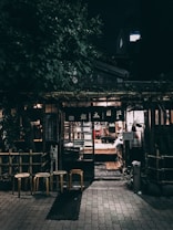 An outdoor night scene depicts a cozy, rustic storefront surrounded by lush greenery. The entrance is illuminated by warm, inviting lights, with three wooden stools positioned in front. The building features traditional wooden elements and a glass door, through which the soft glow of interior lighting can be seen.