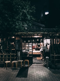An outdoor night scene depicts a cozy, rustic storefront surrounded by lush greenery. The entrance is illuminated by warm, inviting lights, with three wooden stools positioned in front. The building features traditional wooden elements and a glass door, through which the soft glow of interior lighting can be seen.