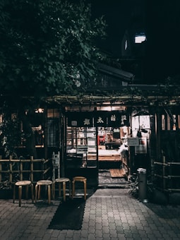 An outdoor night scene depicts a cozy, rustic storefront surrounded by lush greenery. The entrance is illuminated by warm, inviting lights, with three wooden stools positioned in front. The building features traditional wooden elements and a glass door, through which the soft glow of interior lighting can be seen.