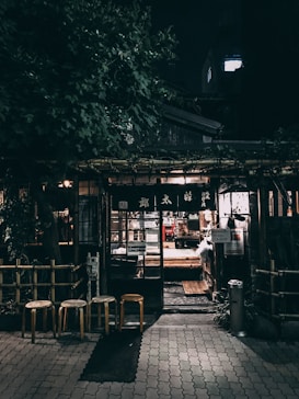 An outdoor night scene depicts a cozy, rustic storefront surrounded by lush greenery. The entrance is illuminated by warm, inviting lights, with three wooden stools positioned in front. The building features traditional wooden elements and a glass door, through which the soft glow of interior lighting can be seen.
