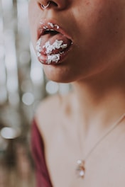 A close-up image of a person's mouth with silver foil applied on the lips and tongue. The individual is wearing a nose ring and a necklace with a pendant. The background features a soft-focus bokeh effect.