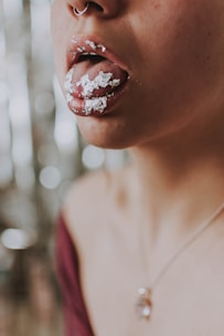A close-up image of a person's mouth with silver foil applied on the lips and tongue. The individual is wearing a nose ring and a necklace with a pendant. The background features a soft-focus bokeh effect.