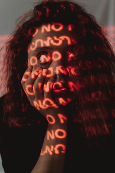 A person's face is obscured by red light projections featuring repeated words, creating an ambiguous and introspective effect. The individual has long, curly hair and a septum piercing, and their hand is placed against their face.