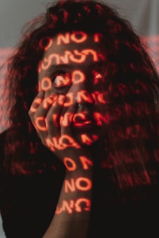 A person's face is obscured by red light projections featuring repeated words, creating an ambiguous and introspective effect. The individual has long, curly hair and a septum piercing, and their hand is placed against their face.