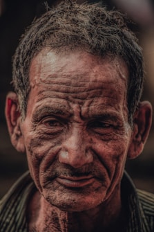 A close-up portrait of an elderly man with deep wrinkles and an expression that appears contemplative. The lighting highlights the texture of his skin, emphasizing the details of his facial features. His hair is short and slightly disheveled.