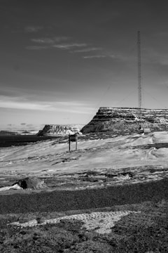 A black and white landscape featuring a tall radio or communication tower rising against a clear sky. In the background, there are rocky hills and expansive flatlands. The terrain appears barren and rugged.
