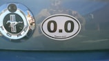 A car trunk features a Ford emblem to the left with a chrome finish and a horse in the center. Next to it, there is a humorous oval sticker reading '0.0 Support Crew', indicating no marathon experience.