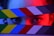 A pair of intense eyes peek through colorful film clapboard slats with vibrant lighting. The lighting creates a dramatic red and blue contrast against the subject's face, which is partially obscured by the clapboard.