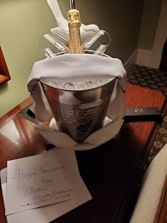 A champagne bottle in an ice bucket, surrounded by a white towel, is displayed on a wooden table. Two glasses flank the bottle. A greeting card with the message 'Happy Anniversary! Enjoy...' is placed next to the bucket. The setting appears elegant and celebratory.