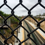 A chain-link fence is prominently featured in the foreground, with train tracks visible in the distance blurred by depth of field. The setting appears to be outdoors, with overcast skies and greenery partially visible on the sides.