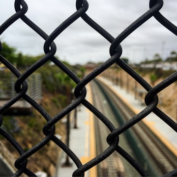 A chain-link fence is prominently featured in the foreground, with train tracks visible in the distance blurred by depth of field. The setting appears to be outdoors, with overcast skies and greenery partially visible on the sides.