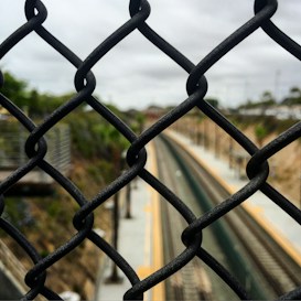 A chain-link fence is prominently featured in the foreground, with train tracks visible in the distance blurred by depth of field. The setting appears to be outdoors, with overcast skies and greenery partially visible on the sides.