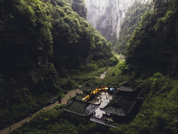A traditional Chinese building complex is nestled at the base of a lush, green mountain gorge. The structures have dark, tiled roofs and are illuminated from within, creating a contrast with the surrounding dense forest. Pathways and small figures of people can be seen around the buildings, indicating activity and exploration in this serene environment.