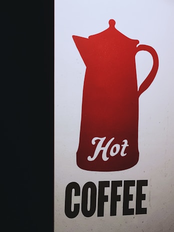A graphic design featuring a red silhouette of a coffee pot with the word 'Hot' written in cursive script. Beneath the pot, the word 'COFFEE' is presented in bold, black uppercase letters on a light background. The design is simple and eye-catching with a strong contrast between the colors.