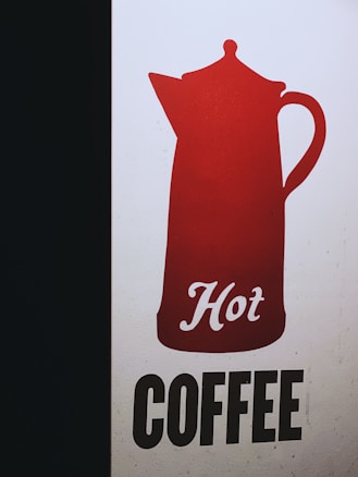 A graphic design featuring a red silhouette of a coffee pot with the word 'Hot' written in cursive script. Beneath the pot, the word 'COFFEE' is presented in bold, black uppercase letters on a light background. The design is simple and eye-catching with a strong contrast between the colors.