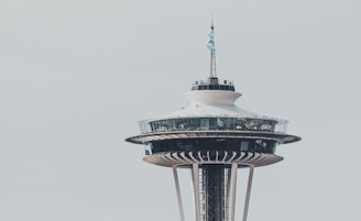 The Space Needle, an iconic observation tower with a flying flag on top, stands against a clear sky. The structure features a wide, circular observation deck with glass windows around it.