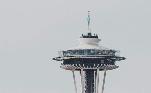 The Space Needle, an iconic observation tower with a flying flag on top, stands against a clear sky. The structure features a wide, circular observation deck with glass windows around it.