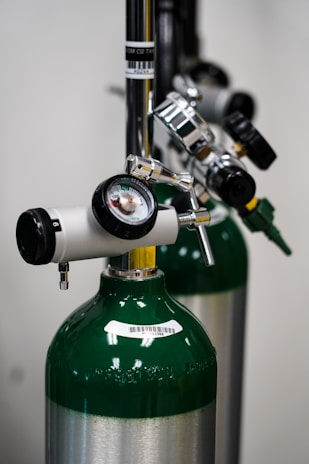 A close-up view of medical oxygen tanks with attached pressure gauges and valves, highlighting the metallic and green elements. The tanks are aligned and feature barcode labels.