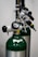 A close-up view of medical oxygen tanks with attached pressure gauges and valves, highlighting the metallic and green elements. The tanks are aligned and feature barcode labels.