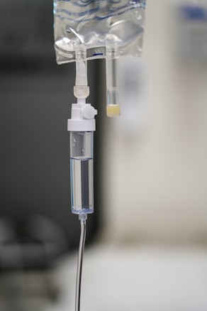 A close-up view of an intravenous (IV) drip bag connected to a tube, containing transparent liquid. The setting appears to be a clinical or hospital environment with blurred background.