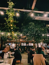 A cozy outdoor dining area with people sitting at wooden tables, surrounded by lush greenery and string lights hanging above. The ambiance is warm and inviting, with a rustic vibe from the brick walls and wooden structures.