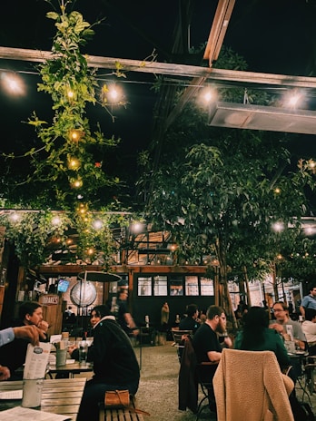 A cozy outdoor dining area with people sitting at wooden tables, surrounded by lush greenery and string lights hanging above. The ambiance is warm and inviting, with a rustic vibe from the brick walls and wooden structures.