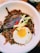 A dish featuring stir-fried noodles topped with a sunny-side-up egg, garnished with sliced beef, sprinkled sesame seeds, fresh cucumbers, and red bell pepper strips. The meal is presented in a large, white bowl with a drink in a glass nearby in the background.