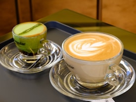 Two glass cups with saucers are placed on a tray. One cup contains a vibrant green matcha drink with a dark layer at the bottom, topped with a line of foamy crema. The other cup holds a cappuccino with an intricate leaf pattern created in the foam, indicating skilled latte art.