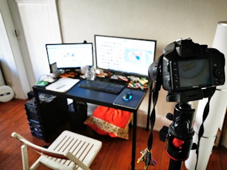 A workspace setup featuring a desk with two computer monitors, a keyboard, mouse, and various office supplies scattered on the surface. A DSLR camera on a tripod is positioned in the foreground, aimed towards the monitors. A white chair is placed next to the desk on a wooden floor.