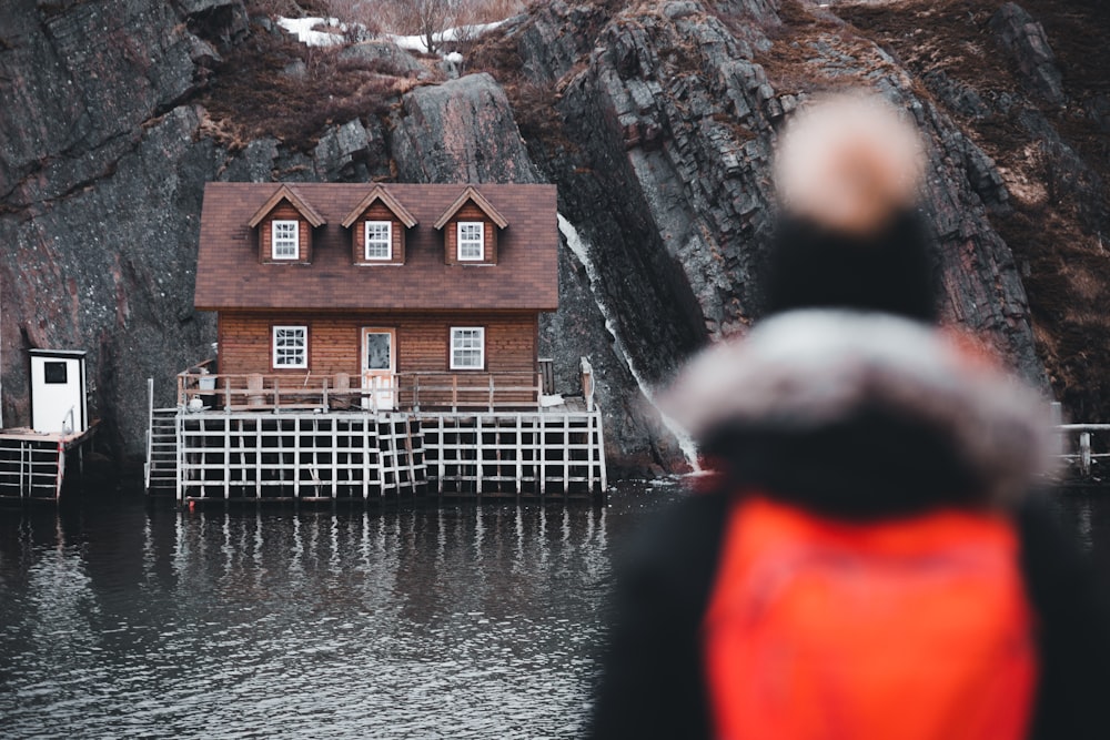 person in red hoodie standing near brown wooden house near body of water during daytime