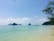 A serene beach with clear turquoise waters gently lapping against the sandy shore. Several longtail boats are floating on the water, and towering limestone islands are visible in the distance under a bright blue sky. Lush greenery is visible on the right side, adding a natural border to the idyllic scene.