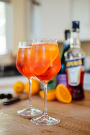 Two wine glasses filled with a vibrant orange cocktail sit on a wooden table. The drinks are garnished with slices of orange. In the background, there are blurred bottles and fresh orange halves, suggesting a casual setting.