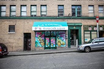 A brick building features a central entrance with a teal awning labeled 'L.A. Lounge Karaoke & Bar.' The doors underneath are decorated with vibrant graffiti, including colorful abstract designs and a horned character with text that reads 'EAT ME.' Two vehicles are parked on the street, and a red lantern-style streetlight is visible.