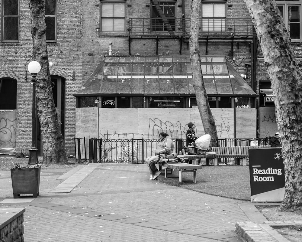 grayscale photo of 2 person sitting on bench near building