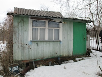 A small, weathered wooden shed with a corrugated metal roof. The exterior is painted in light green and features a window with white frames and a simple green door. The shed is surrounded by snow on the ground. There are bare trees in the background, and some scattered items appear near the base of the shed.