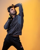 A man strikes a dynamic pose against a warm yellow background, wearing athletic clothing including a black jacket and matching pants, with a gloved hand touching his head.