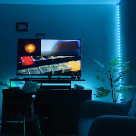 A cozy room illuminated by blue LED lights features a modern entertainment setup with a TV screen displaying a close-up of a control panel. The TV stand holds various electronic components, while a comfortable couch is partially visible. Decorative elements include a potted plant and a small table with a vase of white flowers.