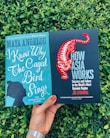 Two books are being held in front of a backdrop of green foliage. The left book is titled 'I Know Why The Caged Bird Sings' by Maya Angelou and has a teal cover with a silhouette of a person's profile. The right book is titled 'How Asia Works' by Joe Studwell and features a black cover with a stylized red tiger.