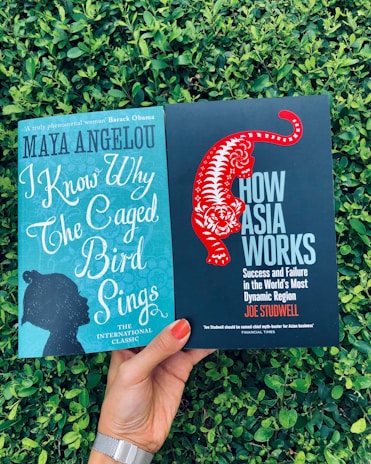 Two books are being held in front of a backdrop of green foliage. The left book is titled 'I Know Why The Caged Bird Sings' by Maya Angelou and has a teal cover with a silhouette of a person's profile. The right book is titled 'How Asia Works' by Joe Studwell and features a black cover with a stylized red tiger.