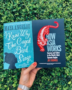 Two books are being held in front of a backdrop of green foliage. The left book is titled 'I Know Why The Caged Bird Sings' by Maya Angelou and has a teal cover with a silhouette of a person's profile. The right book is titled 'How Asia Works' by Joe Studwell and features a black cover with a stylized red tiger.