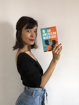 A person with medium-length dark hair, wearing a black top and blue jeans, holds up a copy of the book 'An American Marriage' by Tayari Jones. The background is plain white, and the person faces the camera with a subtle smile.