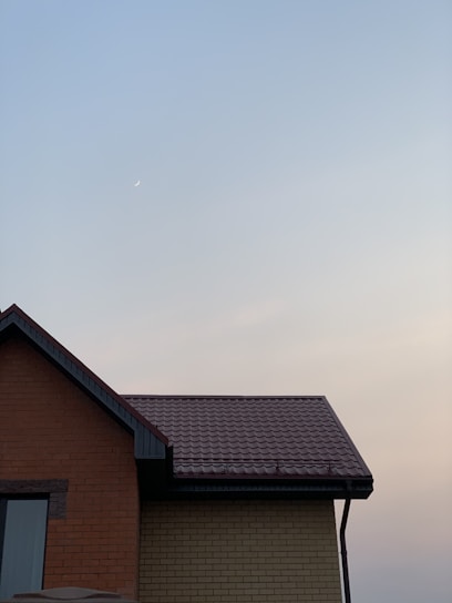 A small crescent moon appears in a clear sky during dusk with a soft gradient from blue to pale orange. The roof of a house with brick and tiled sections is visible at the bottom of the image.