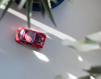 A small red toy car with a Union Jack design on the roof sits on a light-colored surface. The car is partially in the sunlight, with plant leaves creating a soft shadow across the scene.