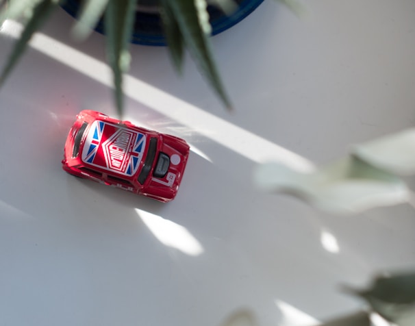 A small red toy car with a Union Jack design on the roof sits on a light-colored surface. The car is partially in the sunlight, with plant leaves creating a soft shadow across the scene.