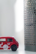 A small toy car in red color is placed on a white surface. The car features detailing with white graphics and miniature decals resembling a Union Jack. Next to the car is a cylindrical object with a perforated metal design. The setting appears to be indoors, with soft, diffused lighting coming from the right side.