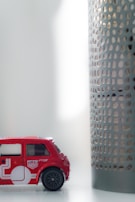 A small toy car in red color is placed on a white surface. The car features detailing with white graphics and miniature decals resembling a Union Jack. Next to the car is a cylindrical object with a perforated metal design. The setting appears to be indoors, with soft, diffused lighting coming from the right side.