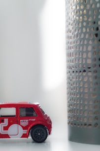 A small toy car in red color is placed on a white surface. The car features detailing with white graphics and miniature decals resembling a Union Jack. Next to the car is a cylindrical object with a perforated metal design. The setting appears to be indoors, with soft, diffused lighting coming from the right side.